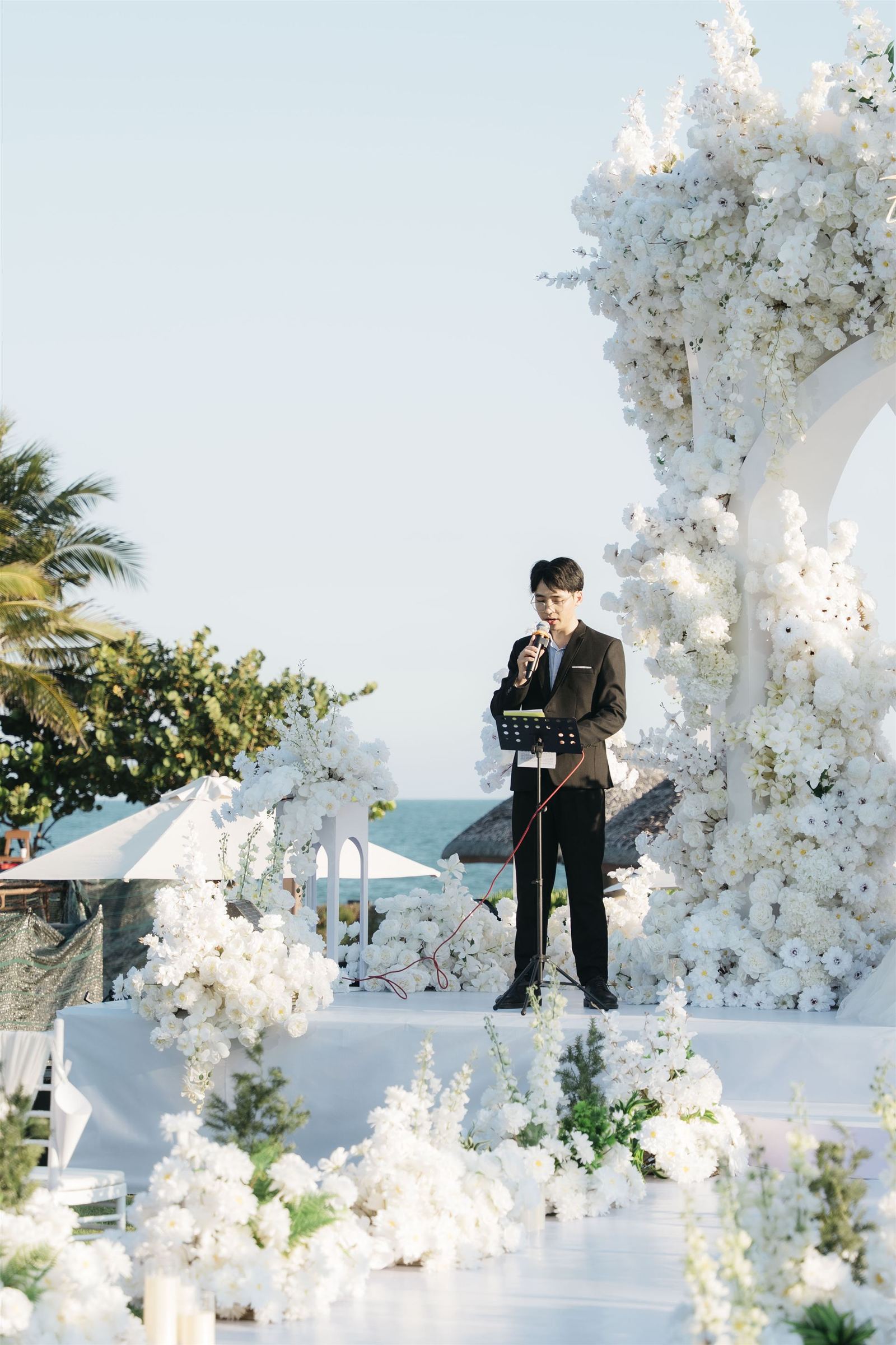 MC Thanh Hiếu cầm mic dẫn lễ cưới ngoài trời bên biển, bên cổng hoa trắng