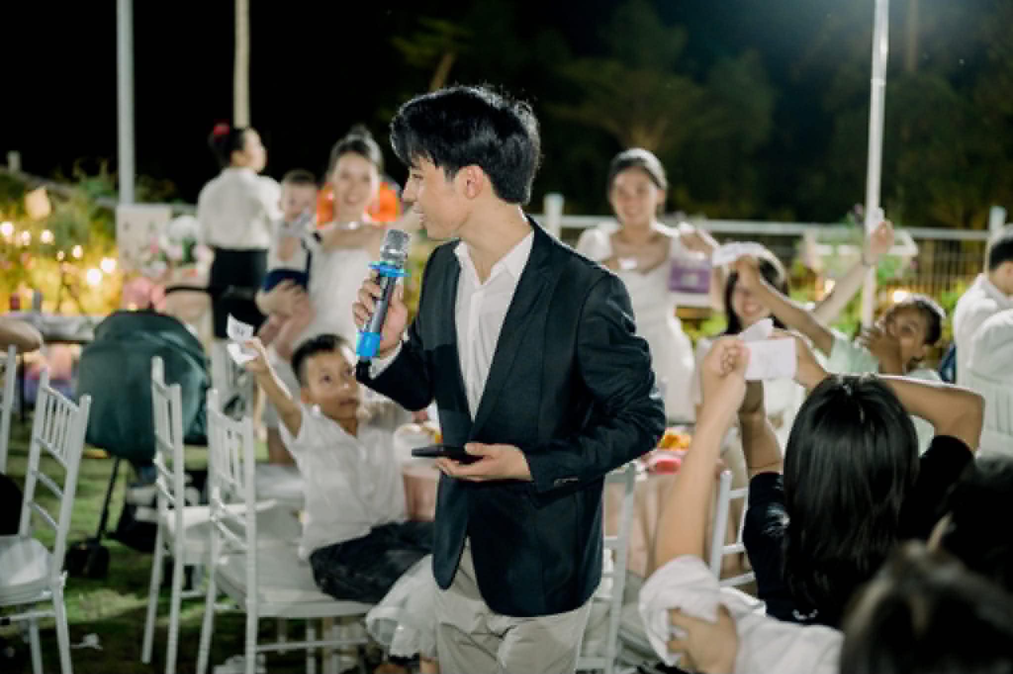 MC Thanh Hiếu cầm mic đi giữa bàn tiệc, khách giơ tay tham gia trò chơi trong tiệc cưới ngoài trời buổi tối