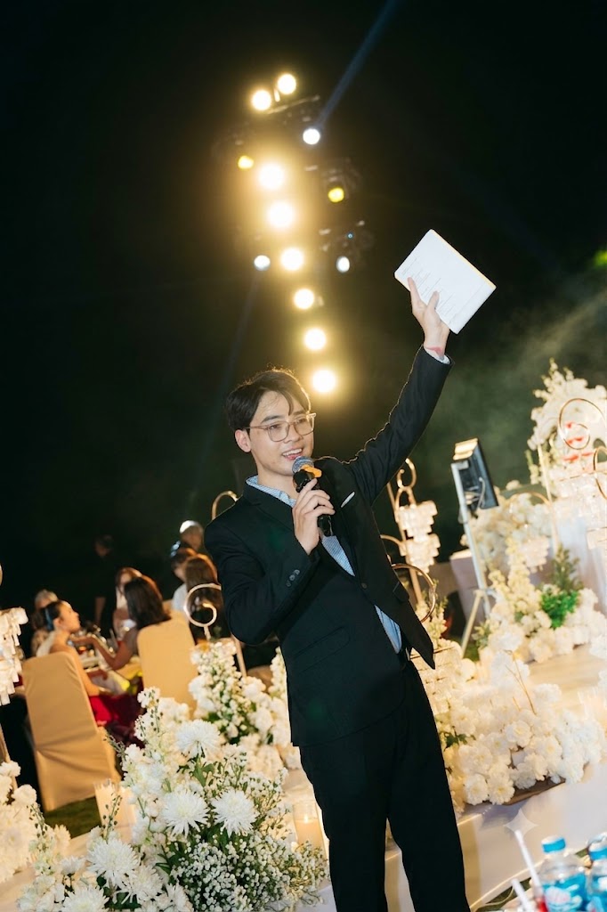 MC Thanh Hiếu giơ tay cầm mic dưới ánh đèn sân khấu trong tiệc cưới
